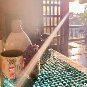 May include: A teal, textured, hand-knitted rug with a mug, vase, and small glass jar with dried flowers. The mug has a colorful design with pink and green accents. A white vase is in the background.