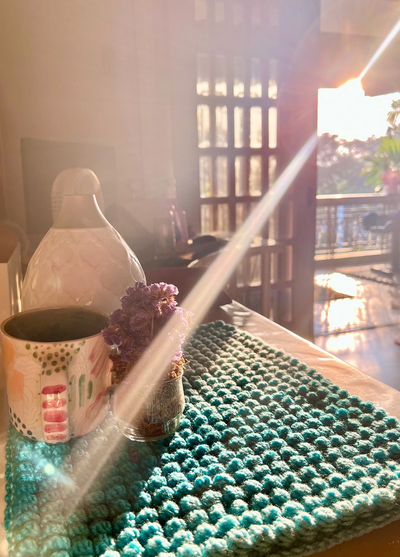 May include: A teal, textured, hand-knitted rug with a mug, vase, and small glass jar with dried flowers. The mug has a colorful design with pink and green accents. A white vase is in the background.