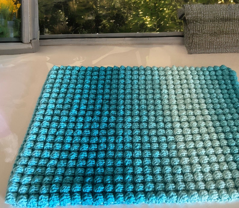 May include: A square, hand-crocheted mat in shades of blue, transitioning from a deep turquoise to a lighter aqua. The textured surface is created by a series of raised, bobble-like stitches, giving it a three-dimensional appearance. The mat is likely used as a decorative item.