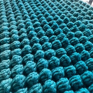 May include: Close-up of a teal-colored, textured knitted fabric. The fabric is densely covered with raised, rounded bobbles, creating a three-dimensional effect. The yarn appears soft and plush, with variations in the teal hue.