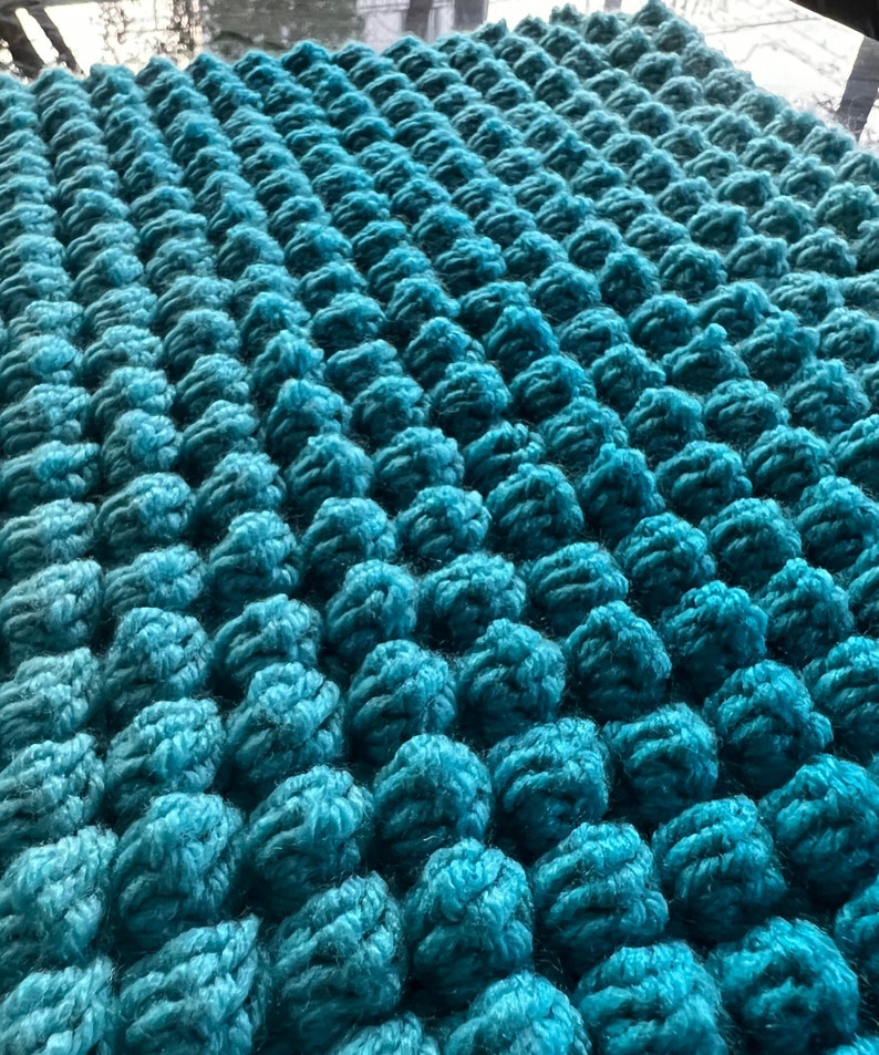 May include: Close-up of a teal-colored, textured knitted fabric. The fabric is densely covered with raised, rounded bobbles, creating a three-dimensional effect. The yarn appears soft and plush, with variations in the teal hue.