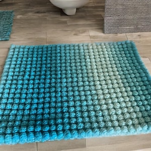 Puede incluir: Una alfombrilla de baño cuadrada con un patrón de textura de puntos de burbuja. La alfombrilla muestra una transición de color degradada, que cambia de un azul verdoso profundo y saturado a la izquierda a un azul aguamarina pálido a la derecha.