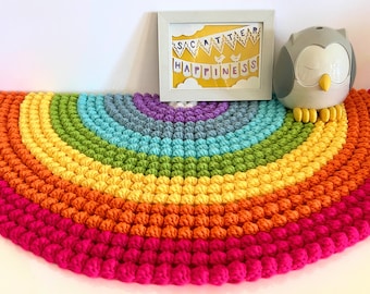 May include: A rainbow crochet rug with a bright pink, orange, yellow, green, blue, purple, and gray color scheme. The rug is in the shape of a half circle and is made of yarn with a textured, puffy look. A white picture frame with the text "Scatter Happiness" is in the background.