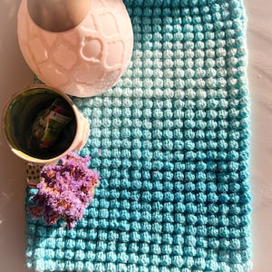 May include: A handmade, square, turquoise and light blue crocheted blanket with a textured, bubble-like pattern. A pink ceramic vase with a decorative pattern and a small green and white cup are next to the blanket. Dried purple flowers are also present.