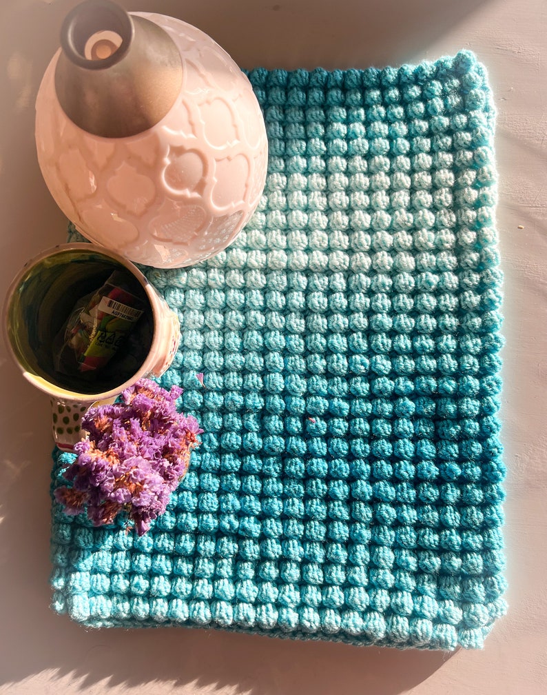 May include: A handmade, square, turquoise and light blue crocheted blanket with a textured, bubble-like pattern. A pink ceramic vase with a decorative pattern and a small green and white cup are next to the blanket. Dried purple flowers are also present.
