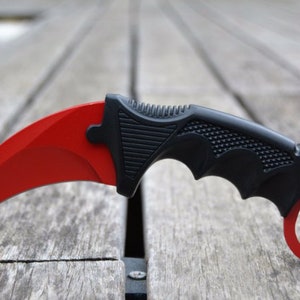 May include: A red and black karambit knife with a curved blade and a finger ring. The knife has a black handle with a textured grip.