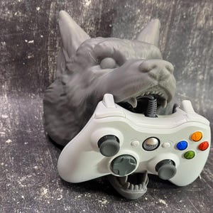 May include: A gray wolf head sculpture with an open mouth holding a white wireless game controller with black buttons and colored buttons. The controller has the word "Voyo" printed on it.
