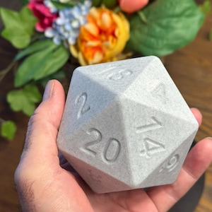 May include: A large, light grey twenty-sided die, with numbers etched on each face. The die is held in a hand, with a blurred floral arrangement in the background. The die is a gaming accessory.