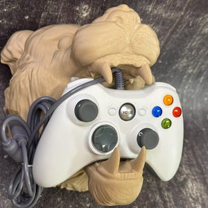 May include: A white Xbox 360 controller with a grey cord is held in the mouth of a 3D printed brown monster figurine. The controller has a black button in the centre and coloured buttons on the right side.