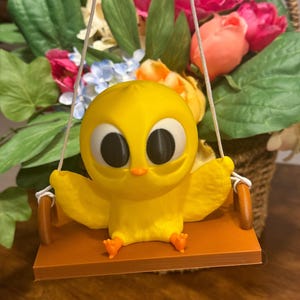 May include: A yellow chick with large black eyes sits on a brown wooden swing. The chick is holding onto the ropes of the swing with its wings. The swing is hanging from a white rope.