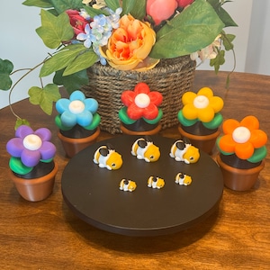 May include: A display of colorful, flower-shaped candles in terracotta pots, alongside miniature guinea pig figurines on a black stand. A basket of vibrant flowers sits in the background, creating a cheerful, decorative arrangement.