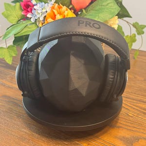 May include: Black over-ear headphones with the word "PRO" printed on the headband, resting on a black geometric stand. The headphones are on a wooden surface, with a colorful floral arrangement in the background.