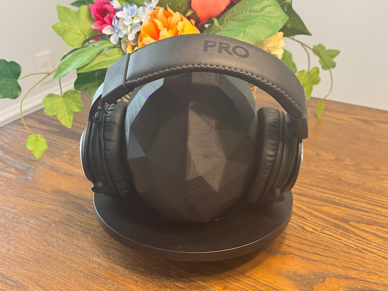 May include: Black over-ear headphones with the word "PRO" printed on the headband, resting on a black geometric stand. The headphones are on a wooden surface, with a colorful floral arrangement in the background.