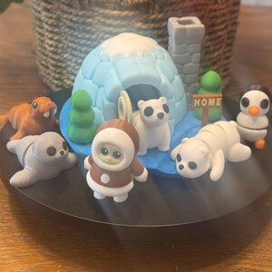 May include: A miniature arctic scene with a white igloo, a brown bear wearing a red and white suit, a white polar bear, a brown seal, a gray seal, a white polar bear, and a black and white penguin. The scene is set on a black base with a blue water feature and a sign that reads "HOME".