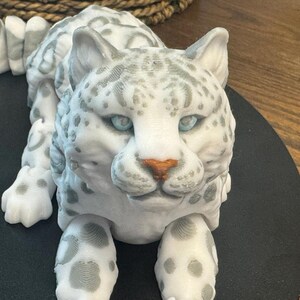 May include: A white snow leopard figurine with gray spots, blue eyes, and a brown nose. The figurine is lying down with its head raised and looking directly at the camera.