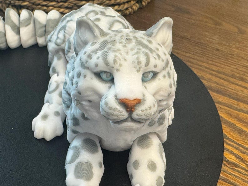 May include: A white snow leopard figurine with gray spots, blue eyes, and a brown nose. The figurine is lying down with its head raised and looking directly at the camera.