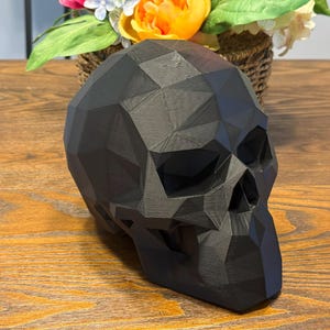 May include: A black, geometric skull sculpture with a faceted design. The skull is made of a matte material and sits on a wooden surface. A basket of flowers is in the background.