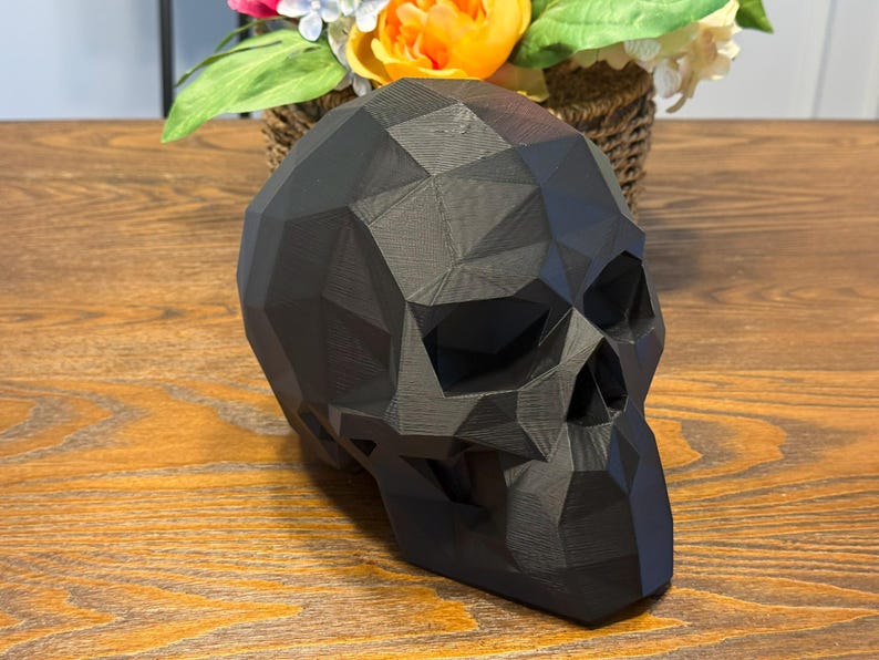 May include: A black, geometric skull sculpture with a faceted design. The skull is made of a matte material and sits on a wooden surface. A basket of flowers is in the background.