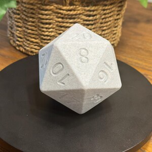 May include: A large, light grey twenty-sided die, with white numbers, sits on a black circular base. The die is a polyhedron with twenty faces. The numbers are clearly visible on the faces of the die. The background includes a woven basket.