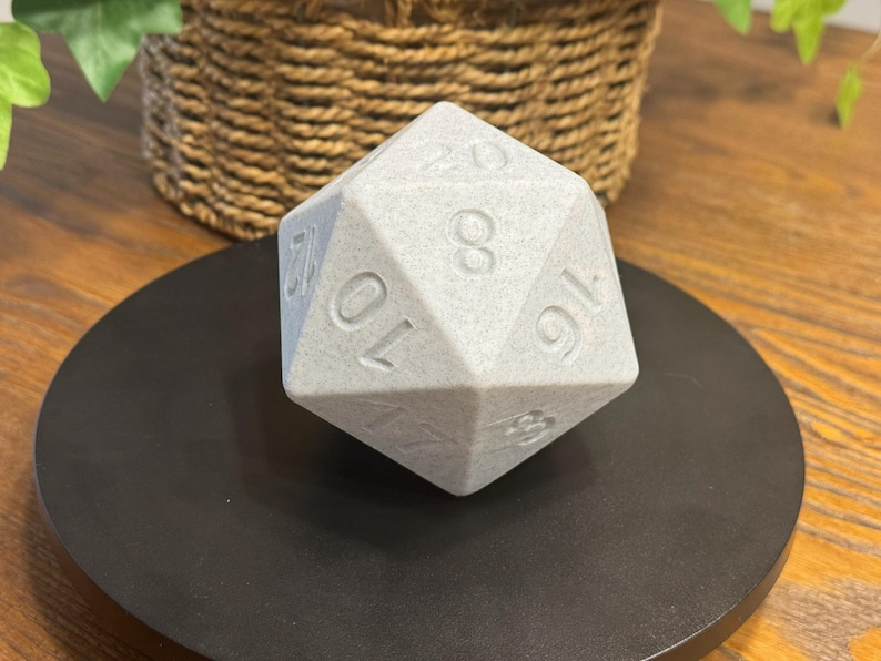 May include: A large, light grey twenty-sided die, with white numbers, sits on a black circular base. The die is a polyhedron with twenty faces. The numbers are clearly visible on the faces of the die. The background includes a woven basket.