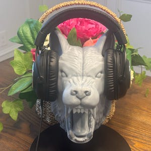 May include: A gray wolf head sculpture with an open mouth, holding black headphones. The sculpture is placed in a woven basket with pink flowers and green foliage. The headphones have a black cord.