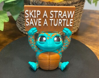 May include: A teal and brown turtle figurine holding a sign that reads "Skip a Straw Save a Turtle".