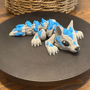 May include: A gray and blue fantasy creature figurine, resembling a wolf or fox, with crystal-like accents. The figurine is lying down on a black circular base. The crystals are a bright blue color.