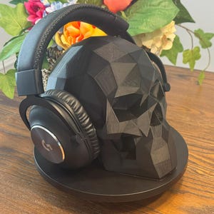 May include: Black over-ear headphones rest on a black geometric skull-shaped stand. The headphones have a silver logo on the ear cup. The stand is on a wooden surface, with colorful flowers in the background.