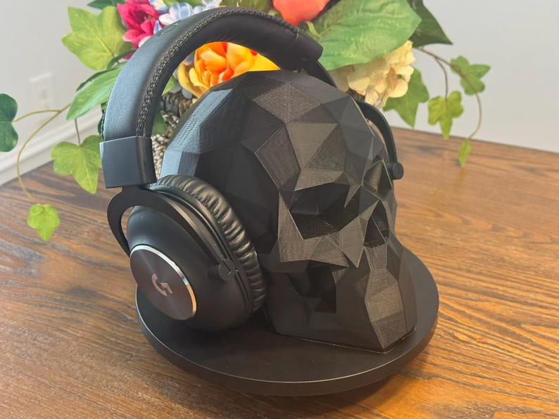 May include: Black over-ear headphones rest on a black geometric skull-shaped stand. The headphones have a silver logo on the ear cup. The stand is on a wooden surface, with colorful flowers in the background.