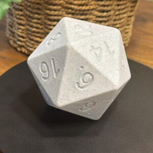 May include: A large, light grey, twenty-sided die with engraved numbers. The die is resting on a black surface, with a woven basket and greenery in the background. The numbers are clearly visible on the die's faces.