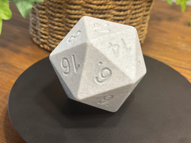 May include: A large, light grey, twenty-sided die with engraved numbers. The die is resting on a black surface, with a woven basket and greenery in the background. The numbers are clearly visible on the die's faces.