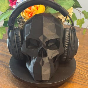 May include: A black skull-shaped headphone stand with a geometric design. The stand holds a pair of black over-ear headphones. The stand is on a round black base, with flowers in the background.