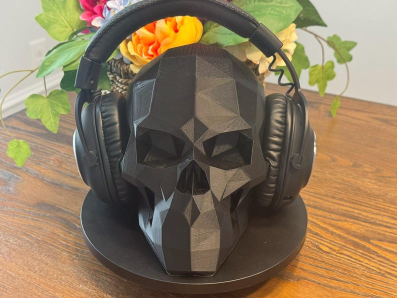 May include: A black skull-shaped headphone stand with a geometric design. The stand holds a pair of black over-ear headphones. The stand is on a round black base, with flowers in the background.
