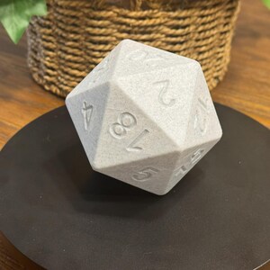 May include: A large, grey, twenty-sided die with engraved numbers. The die is resting on a black, circular surface. The background includes a woven basket and greenery, suggesting a tabletop setting. Ideal for tabletop gaming.