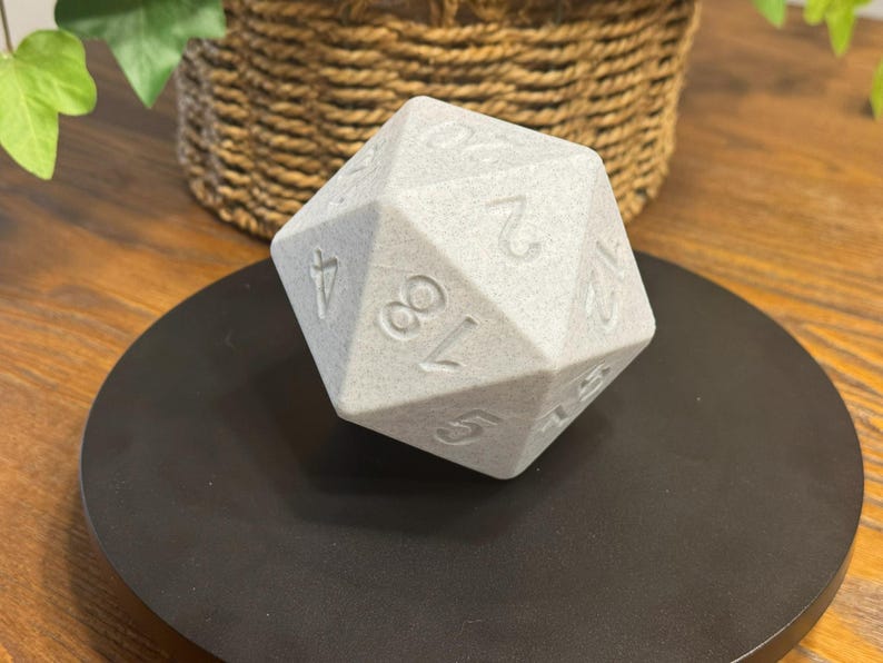 May include: A large, grey, twenty-sided die with engraved numbers. The die is resting on a black, circular surface. The background includes a woven basket and greenery, suggesting a tabletop setting. Ideal for tabletop gaming.