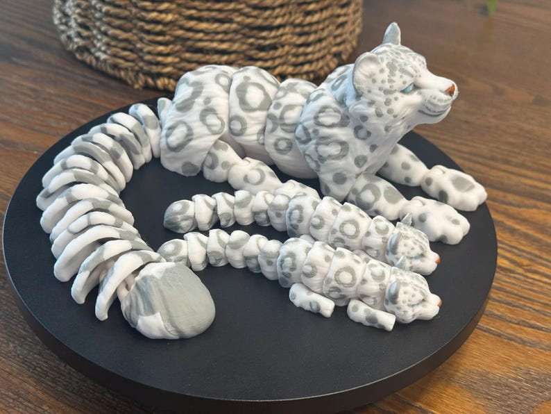 May include: A white and gray snow leopard figurine with a curled tail and two smaller snow leopard figurines. The figurines are made of a smooth, white material with gray spots.