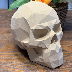 May include: A beige, geometric skull sculpture with a faceted design. The skull is positioned on a wooden surface, with a woven basket of flowers in the background. The sculpture has a modern, artistic aesthetic.