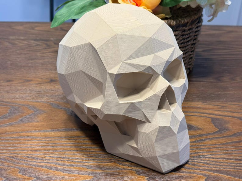 May include: A beige, geometric skull sculpture with a faceted design. The skull is positioned on a wooden surface, with a woven basket of flowers in the background. The sculpture has a modern, artistic aesthetic.