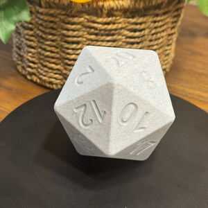 May include: A light grey, multi-sided die with engraved numbers, resting on a black circular surface. The die is a polyhedron, likely used in tabletop games. The numbers are clearly visible, indicating its function for random number generation.
