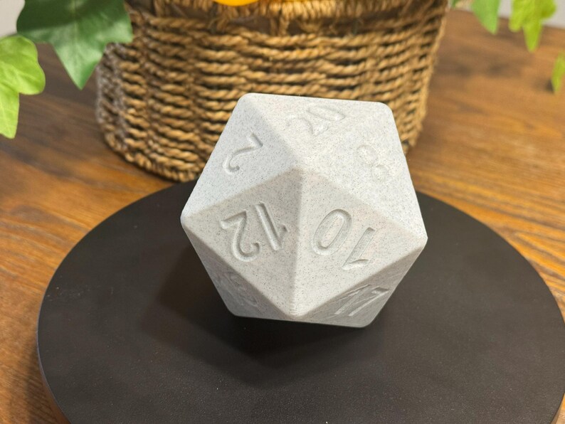 May include: A light grey, multi-sided die with engraved numbers, resting on a black circular surface. The die is a polyhedron, likely used in tabletop games. The numbers are clearly visible, indicating its function for random number generation.