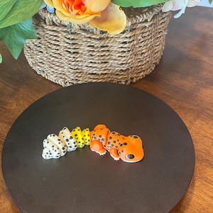 May include: A small, orange, yellow, and white gecko figurine with black spots. The gecko is posed in a curled position on a black circular surface.