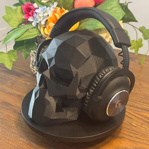 May include: A black, geometric skull-shaped headphone stand with a pair of black headphones resting on it. The stand is on a round black base. The background includes a floral arrangement with colorful flowers.