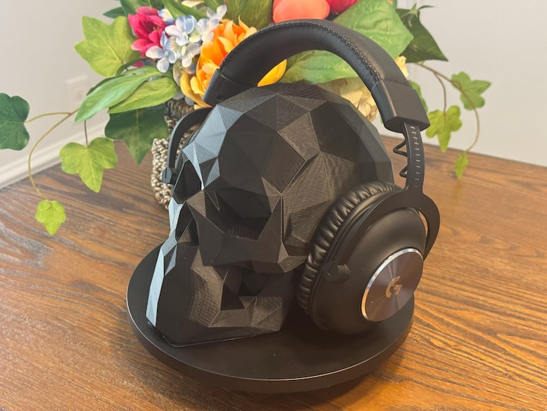 May include: A black, geometric skull-shaped headphone stand with a pair of black headphones resting on it. The stand is on a round black base. The background includes a floral arrangement with colorful flowers.