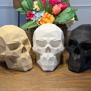 May include: Three geometric skull sculptures in beige, white, and black are displayed on a wooden surface. The skulls have a faceted design and are positioned in front of a basket of colorful flowers, creating a contrasting decorative arrangement.