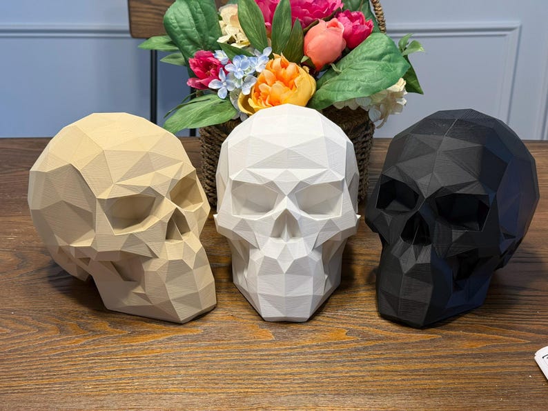 May include: Three geometric skull sculptures in beige, white, and black are displayed on a wooden surface. The skulls have a faceted design and are positioned in front of a basket of colorful flowers, creating a contrasting decorative arrangement.