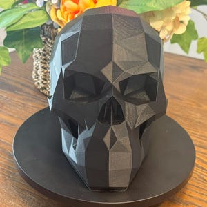 May include: A black, geometric skull sculpture with a faceted design. The skull is displayed on a black circular base, with a blurred background of flowers and greenery. The sculpture is a decorative item.