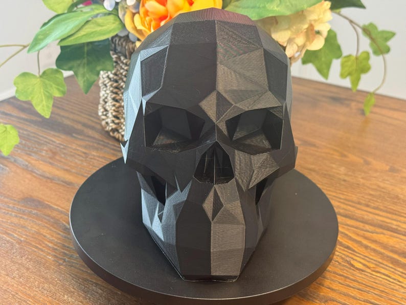 May include: A black, geometric skull sculpture with a faceted design. The skull is displayed on a black circular base, with a blurred background of flowers and greenery. The sculpture is a decorative item.