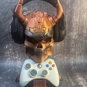 May include: A gold and brown skull-shaped gaming headset stand with a white video game controller. The stand has horns and a black headset is resting on the horns.