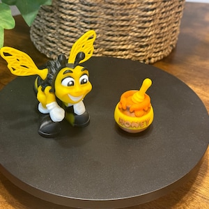 May include: A yellow and black bee figurine with a smiling face and white gloves, standing next to a small yellow pot of honey with the word "honey" printed on it.