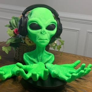 May include: A green alien bust wearing black headphones with outstretched green hands. The alien has large eyes and a serious expression. The bust is on a black circular base.